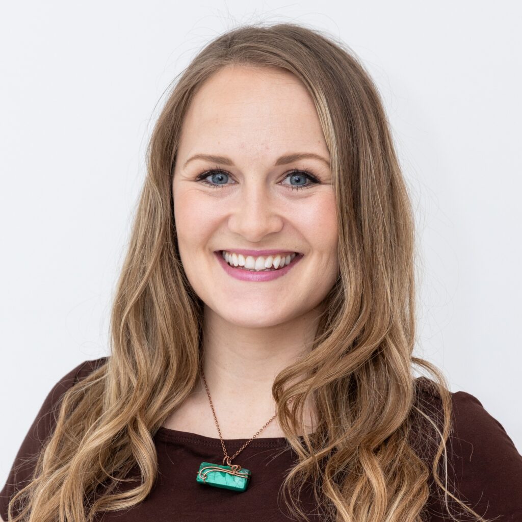 Close-up of a smiling woman with long light brown hair, blue eyes, wearing a turquoise pendant necklace on a copper chain.